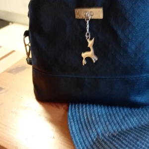 Svart axelväska med hjortdetalj - Snygg svart axelväska i syntetmaterial med quiltad yta och en charmig hjortdetalj i metall. Väskan har en praktisk dragkedja upptill och en justerbar axelrem. Perfekt för att bära dina dagliga nödvändigheter med stil.