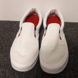 Vita slip-on sneakers från Tommy Hilfiger - Snygga vita slip-on sneakers från Tommy Hilfiger med röd insida och elastiska band i blått och rött. Perfekta för en stilren och bekväm look.