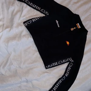 Äkta Svart tröja med dragkedja från Ellesse - Snygg och trendig Ellesse tröja med halvlång dragkedja. Tröjan är svart med vita Ellesse-loggor längs ärmen. Den har en liten Ellesse-logga på bröstet. Perfekt för både vardag och träning. Använd 2 gånger.