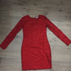 Röd spetsklänning - Snygg röd spetsklänning med långärmad design och blommigt mönster. Klänningen har en elegant öppen ryggdetalj och är perfekt för speciella tillfällen. Den är tillverkad i ett vackert spetsmaterial som ger en lyxig känsla.