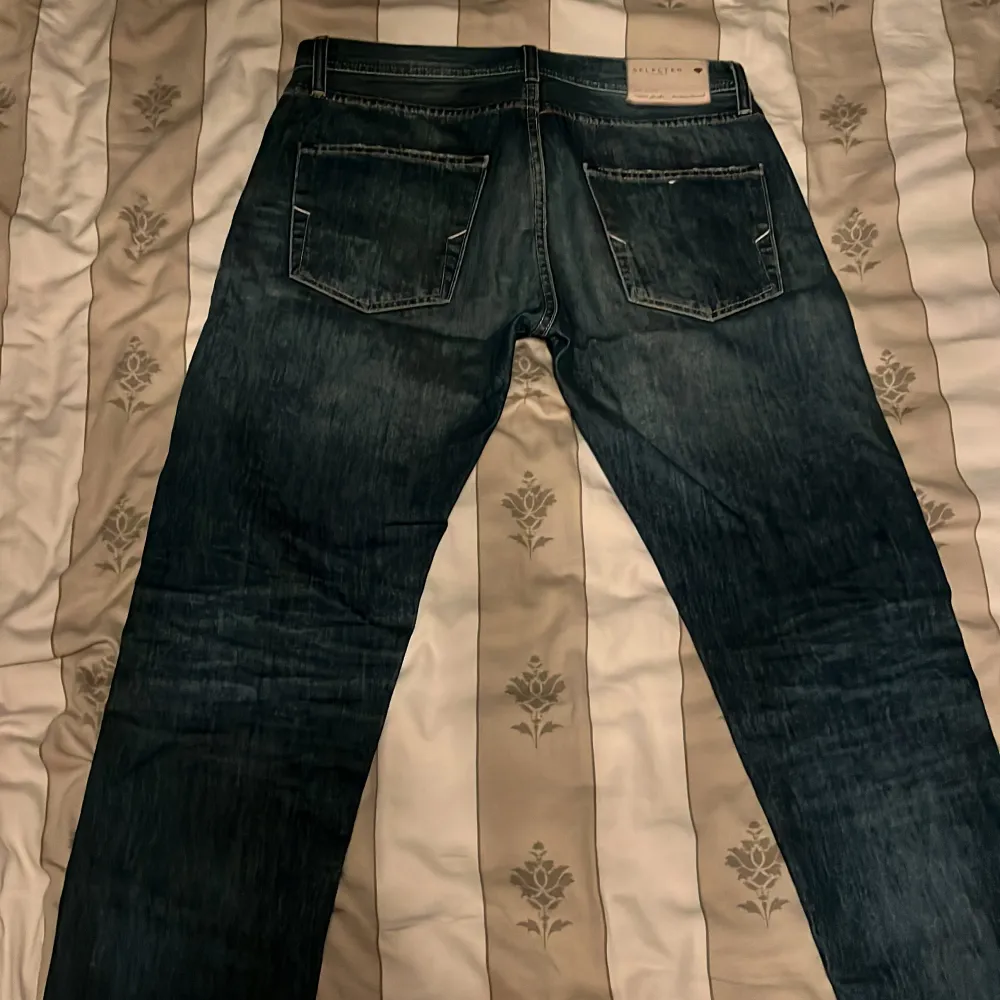 Snygga mörkblå jeans från Selected Homme med en klassisk femficksdesign. Jeansen har en diskret tvättad look och är dekorerade med små färgdetaljer vid linningen. Perfekta för en stilren vardagslook. Storlek 29/34. Nypris 800. Farkut & Housut.
