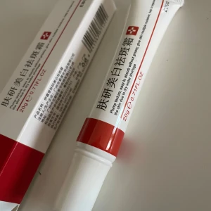 Whitning creme för fräknar/fläckar  - En vit tub med röd text och detaljer, innehållande 20g produkt. Förpackningen har en enkel och stilren design med en röd rand nära öppningen.