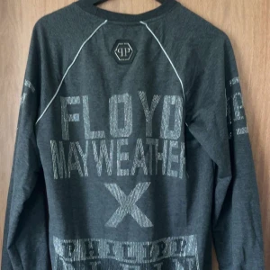 Grå långärmad tröja från Philipp Plein limitid edition - Snygg grå långärmad tröja från Philipp Plein med texten 'Floyd Mayweather' i glittriga bokstäver. Tröjan har en sportig look med vita sömmar och är perfekt för fans av boxning.