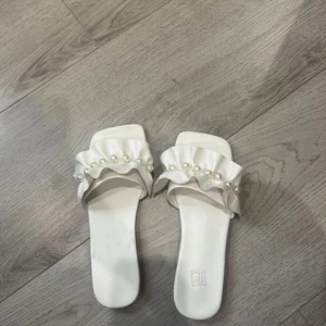 Vita sandaler med pärldetaljer - Eleganta vita sandaler med volang och pärldetaljer. Perfekta för en stilren look. Sandalerna har en fyrkantig tå och en platt sula för bekväm passform.