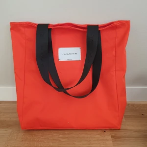 Orange shoppingväska från La Manufacture - Säljer en snygg orange shoppingväska från La Manufacture. Väskan har svarta handtag och en stilren design med en broderad logotyp på ena sidan. Perfekt för att bära med sig allt man behöver under dagen. 37×34cm