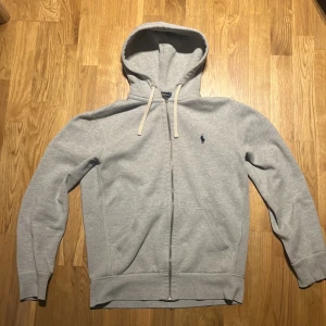 Grå hoodie från Polo Ralph Lauren - Säljer en stilren grå hoodie från Polo Ralph Lauren. Den har en dragkedja framtill och en klassisk broderad logga på bröstet. Perfekt för en avslappnad look med sina långa ärmar och bekväma passform.