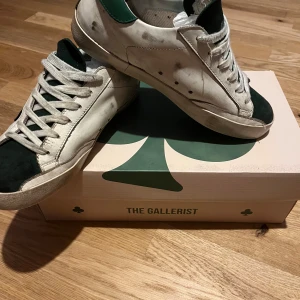 Vita och gröna sneakers från The Gallerist - Snygga vita sneakers med gröna detaljer och svart klövermotiv från The Gallerist. Skorna har snörning och en robust sula. Perfekta för en stilren look. Dom är använda men bra skick sulan är lite sliten 