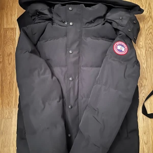  Canada Goose - Säljer en svart dunjacka från Canada Goose med deras ikoniska märke på ärmen. Jackan har en stilren design med knappar och dragkedja framtill. Perfekt för kalla vinterdagar.