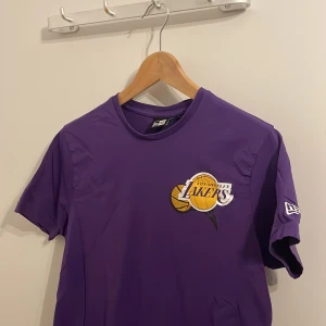 Lila Los Angeles Lakers t-shirt från New Era - Säljer en lila t-shirt från New Era med Los Angeles Lakers-logga på bröstet och ryggen. T-shirten är kortärmad och har en klassisk rund halsringning. Perfekt för alla Lakers-fans! 🏀Kom i fel storlek så är ej använd! 