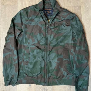 Camouflage vindjacka från Woolrich - Säljer en cool camouflage vindjacka från Woolrich. Jackan har en dragkedja framtill och två praktiska fickor. Perfekt för en avslappnad stil och skyddar mot vinden. Den har en klassisk krage och elastiska muddar vid ärmslut och nederkant.
