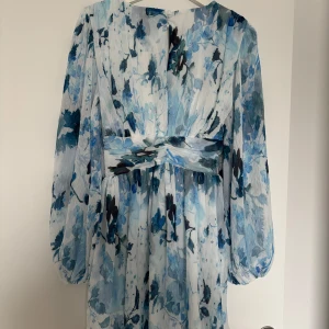 Nu Blommig blå klänning från NA-KD - Säljer en vacker blå blommig klänning med långa ärmar och omlott-design. Klänningen har en flowy känsla och är perfekt för sommarens festligheter. Den har en v-ringad hals och ett fint mönster i olika blå nyanser.
