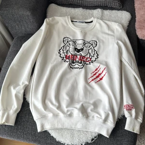 Vit sweatshirt från Kenzo med tigerbroderi - Säljer en vit sweatshirt från Kenzo med ett coolt tigerbroderi i svart och rött på framsidan. Tröjan har långa ärmar och ribbade muddar vid ärmslut och nederkant. Perfekt för en stilren look! Ny