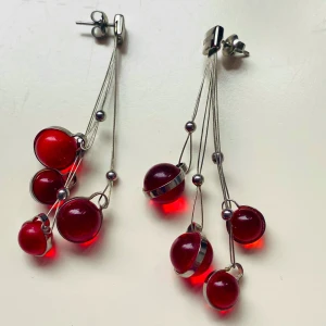 Röda örhängen från Storm - Säljer ett par coola röda örhängen från Storm. De har en unik design med flera röda kulor som hänger i olika längder på tunna silverfärgade trådar. Perfekta för att ge en färgklick till din outfit!