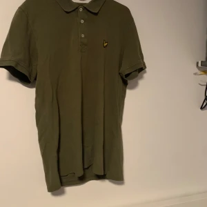 Olivgrön pikétröja från Lyle & Scott - Säljer en snygg olivgrön pikétröja från Lyle & Scott. Den har en klassisk krage med knappar och det ikoniska Lyle & Scott-logotypen på bröstet. Perfekt för en avslappnad men stilren look! Står L men passar som en S