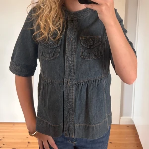 Jeansjacka med kort ärm från Indiska - Säljer en snygg jeansjacka/topp från Indiska med korta ärmar och knappar framtill. Jackan/toppen har två bröstfickor med knappdetaljer och en fin broderad detalj på insidan. Perfekt för hösten🍁🌬️🍂💙