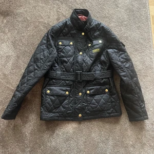 Jacka Barbour (Ungdom) 🌸 - Barbour Internationals klassiska diamant-quiltade jacka i slimmad design med bälte i midjan, trattkrage med halsrem och fem fickor varav en på ärmen. Storlek XL motsvarar XS i dam 