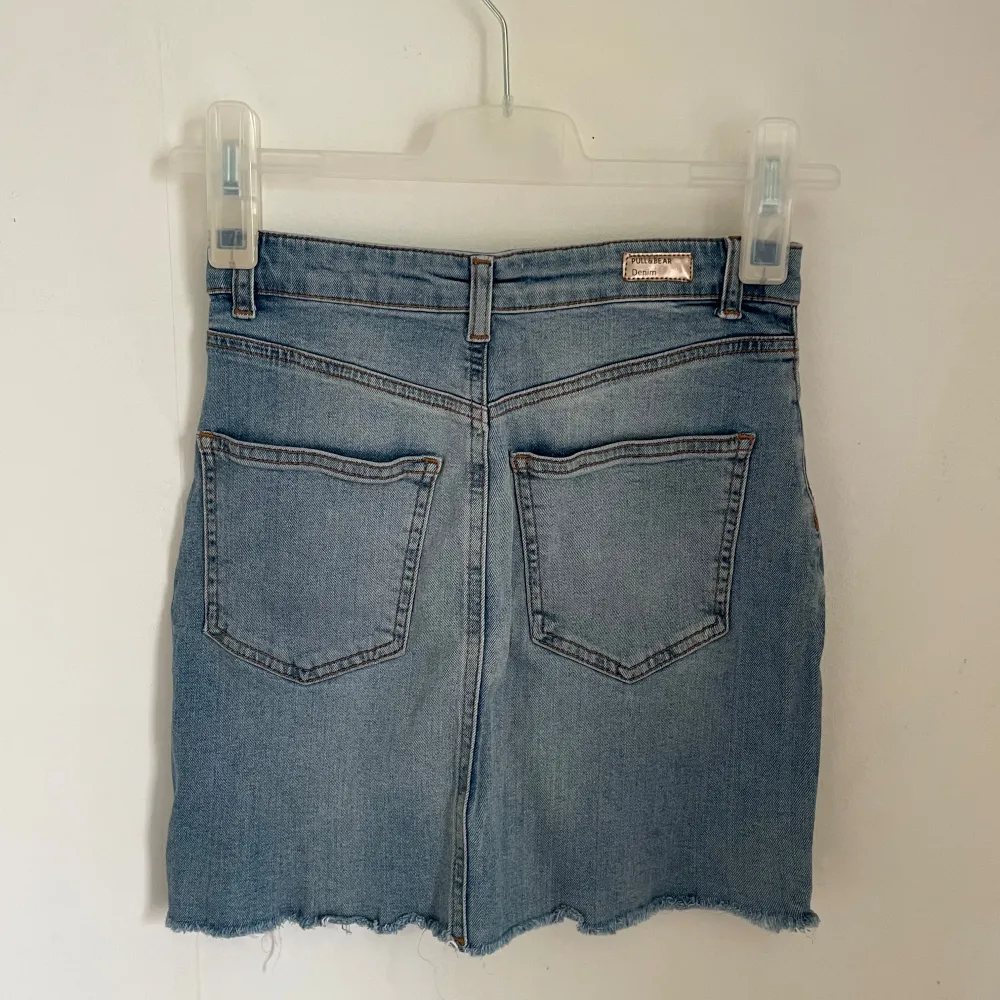 Säljer en snygg jeanskjol från Pull&Bear i storlek S. Kjolen är i en klassisk blå denimfärg och har en cool knappdetalj framtill. Den har en rå kant nertill och en liten slits som ger den en avslappnad look. Perfekt för både vardag och fest!. Kjolar.