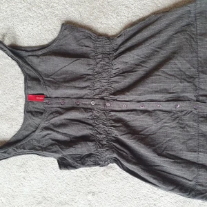 Grå topp från Vero Moda - Säljer en snygg topp från Vero Moda. Den är ärmlös och har knappar hela vägen ner framtill. Kritstreckrandig och smockad detalj i midjan. Strl M.