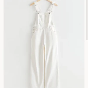 Vit byxdress med hängslen - Säljer en superfin vit byxdress med justerbara hängslen och knappar på sidorna. Den har en avslappnad passform och är perfekt för en stilren look. Materialet känns mjukt och bekvämt, vilket gör den idealisk för både vardag och fest.