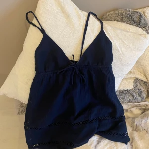 Mörkblå topp från Gina Tricot - Säljer en snygg mörkblå topp från Gina Tricot. Den är ärmlös med smala axelband och har en söt knytning framtill. Perfekt för sommardagar eller en avslappnad festkväll. Toppen har fina spetsdetaljer längst ner som ger den en extra touch.