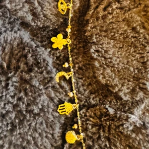 Guldarmband med hängande berlocker - Säljer ett superfint guldarmband från pilgrim med flera hängande berlocker. Armbandet har detaljer som en blomma, en måne, en hand, ett öga och en snäcka. Perfekt för att ge en bohemisk touch till din outfit. Passar både till vardags och fest!