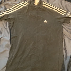 Svart t-shirt från Adidas - Säljer en svart t-shirt från Adidas med de klassiska tre ränderna i vitt längs ärmarna. T-shirten har en normal passform och är gjord i mjuk bomull. Perfekt för både träning och vardag. Adidas-loggan finns både på bröstet och ärmen. Skicket är bra och den är redo för en ny ägare!