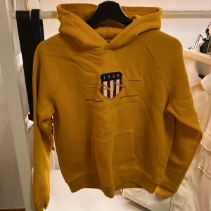 Gul hoodie från GANT - Säljer en snygg gul hoodie från GANT med ett coolt emblem på bröstet. Den har en klassisk känguruficka och är perfekt för höst och vinter. Superbekväm och stilren design!