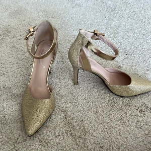 Guldiga klackskor från Ling Mei - Säljer ett par glittriga guldiga klackskor från Ling Mei. De har en spetsig tå och en elegant rem med spänne runt ankeln. Perfekta för festliga tillfällen!
