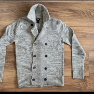 Grå stickad kofta från H&M - Säljer en stilren grå stickad kofta från H&M med dubbelknäppning och ribbade detaljer vid krage och ärmslut. Perfekt för kyligare dagar och ger en klassisk look. Passar både till vardags och mer uppklädda tillfällen.
