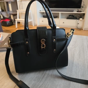 Svart handväska från Steve Madden - Snygg svart handväska från Steve Madden med eleganta gulddetaljer och ett stilrent monogram på framsidan. Väskan har både handtag och en avtagbar axelrem för flexibilitet. 
