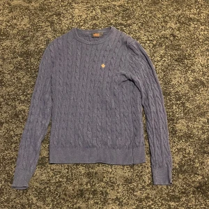 Grå stickad tröja från Woolrich - Säljer en stilren grå stickad tröja från Woolrich med kabelstickat mönster och en liten broderad detalj i gult på bröstet. Tröjan har långa ärmar och ribbade muddar vid ärmslut och nederkant. Perfekt för kyligare dagar!