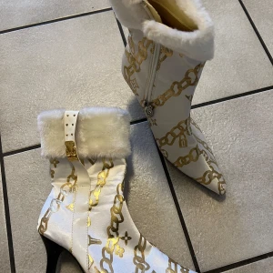 Vita stövletter med gulddetaljer från Louis Vuitton - Eleganta vita stövletter med guldfärgade kedjemönster och fluffig pälsdetalj vid skaftet. Skorna har en spetsig tå och klack, samt en dekorativ spänne. Perfekta för att göra ett statement.