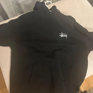 Svart hoodie från Stüssy - Säljer en svart hoodie från Stüssy med deras ikoniska logga i vitt på bröstet och ryggen. Tröjan har en stor ficka framtill och en bekväm huva med dragsko. Perfekt för en avslappnad stil. Fräsch och som ny, inga tecken på användning 