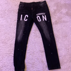 Svarta jeansbyxor från Dsquared2 - Snygga svarta jeansbyxor från Dsquared2 med texten 'ICON' tryckt i vitt på framsidan. Byxorna har en modern look med slitningar och en cool detalj i form av en ring baktill. Perfekta för en trendig stil.