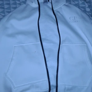Vit hoodie från C.P. Company - Säljer en stilren vit hoodie från C.P. Company. Den har en dragkedja framtill och två praktiska fickor. Perfekt för en avslappnad look.