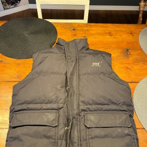 Svart dunväst från Helly Hansen STRL:S - Säljer en svart dunväst från Helly Hansen. Västen har en hög krage och stängs med både dragkedja och knappar. Två stora fickor framtill ger extra förvaring. Perfekt för kyligare dagar. Sitter lite för liten på mig som är 180cm lång och väger 70kg.