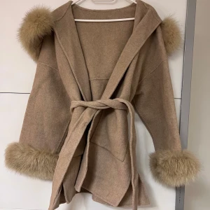 Beige kappa med pälsdetaljer - Snygg brun/beige kappa med pälsdetaljer på huva och ärmslut. Kappan har en knytning i midjan för en elegant look. Perfekt för kyliga dagar när du vill hålla stilen.