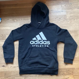Svart hoodie från Adidas 152 - Säljer en svart hoodie från Adidas med stor logga på framsidan.  Storlek 152!! Tröjan har en klassisk känguruficka och en bekväm huva. Perfekt för en sportig look eller avslappnade dagar. 🖤