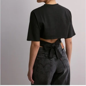 Svart knytbar topp från Nelly - Säljer en svart topp från Nelly med knytband i ryggen. Toppen har korta ärmar och en croppad design som ger en modern look. Perfekt för att styla med jeans eller kjol.