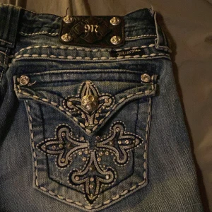 Blå jeans från Miss Me - Snygga blå jeans från Miss Me med unika detaljer på bakfickorna. Jeansen har en dekorativ broderad design med glittriga stenar och en bootcut-stil. Perfekt för dig som vill ha något extra i garderoben!