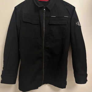 Svart jacka från Calvin Klein - Snygg svart jacka från Calvin Klein med dragkedja framtill och två bröstfickor. Jackan har långa ärmar och en stilren design med en liten logga på ärmen. Perfekt för en cool och avslappnad look.