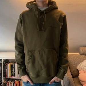 Olivgrön Samsoe hoodie - Säljer en olivgrön hoodie med känguruficka och justerbar huva. Perfekt för en avslappnad stil. Passar bra till jeans eller joggers. Personen på bilden är 170 och hör av er vid frågor och funderingar! Priset ej hugget i sten!