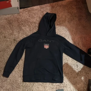Mörkblå hoodie från GANT - Säljer en snygg mörkblå hoodie från GANT med en känguruficka och ett stilrent tryck på bröstet. Perfekt för en avslappnad stil. Huvan ger extra komfort och värme.