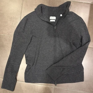 Grå merinoulltröja från These Glory Days - Säljer en stilren grå tröja i merinoull från These Glory Days. Tröjan har en praktisk dragkedja och långa ärmar, perfekt för kyligare dagar. Den är mjuk och bekväm med en klassisk design som passar till det mesta. Perfekt för dig som vill ha både stil och komfort. Storlek M men passar S