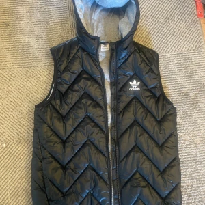 Svart dunväst från Adidas - Snygg svart dunväst från Adidas med dragkedja och huva. Västen har ett quiltat mönster och en grå insida. Perfekt för kyligare dagar när du vill hålla stilen.