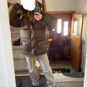 Brun dunjacka från Fjällräven - Säljer en brun dunjacka från Fjällräven med gula detaljer. Jackan har en pufferdesign med dragkedja och knappar framtill. Den är utrustad med en huva och praktiska fickor med knappar. Perfekt för kyliga dagar. Har tyvärr fått fläckar på ryggen och höger fram ficka, men är inte synliga. Hör av er om frågor!💕Inget pis är spikat. Kan gå ner i pris🫶