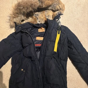 Mörkblå dunjacka med pälsdetaljer från Parajumpers - Säljer en mörkblå dunjacka från Parajumpers med pälsdetaljer. Jackan har en gul dragkedja och flera praktiska fickor och är i storlek small Perfekt för kalla vinterdagar. Inget kvitto kvar därav det billiga pris! 