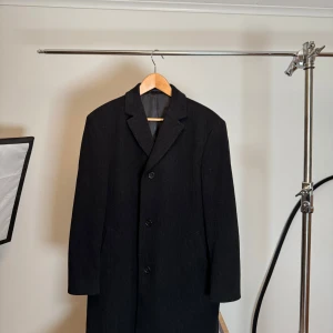 Klassisk kappa från Hugo Boss - Svart kappa från Hugo Boss med klassisk design. Kavajen har en stilren passform och är tillverkad i en ullblandning för en lyxig känsla. Perfekt för formella tillfällen.