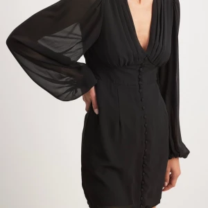 Svart klänning från NA-KD - Elegant svart klänning från NA-KD med djup v-ringning och långa, transparenta ärmar. Klänningen har knappar framtill som ger en stilren look. Perfekt för en kväll ute eller en speciell tillställning.