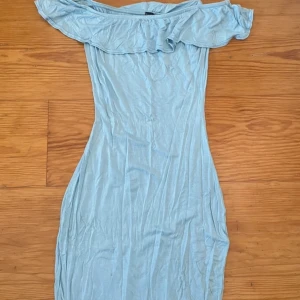 Ljusblå offshoulder klänning - Säljer en ljusblå offshoulder klänning med volangdetalj. Klänningen har en tight passform och är kort. Lite färgmissningar på något ställe.